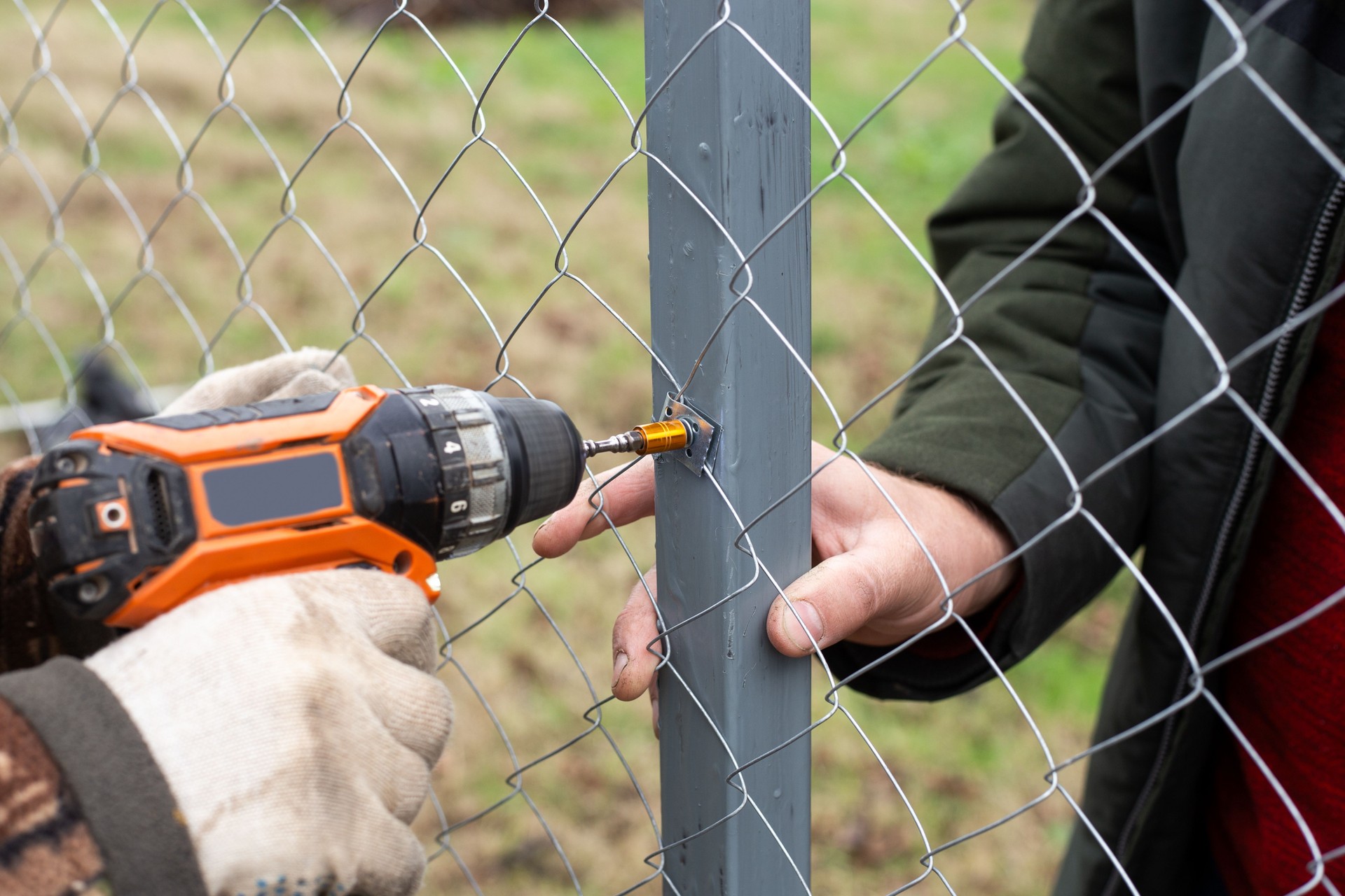 A man is building a fence at his dacha. Using an electric drill, he screws a metal mesh chain-link to the metal profile